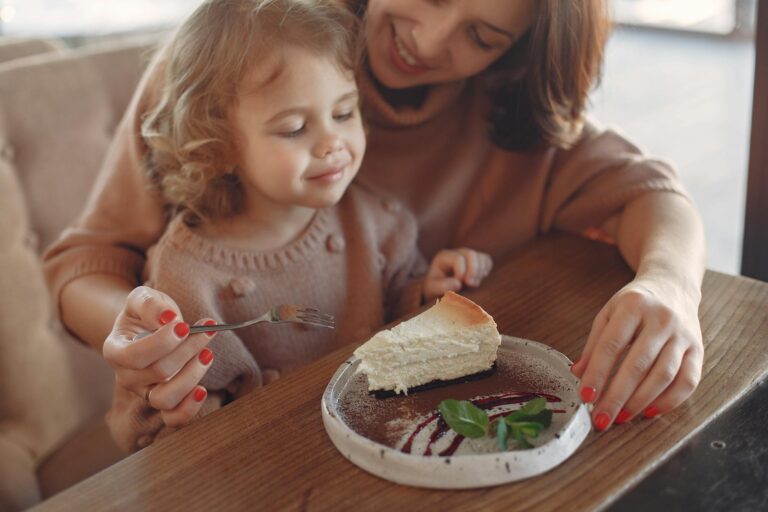 Crop happy mother in casual clothes sitting at table and eating piece of cake with little girl while spending time at cafeteria together