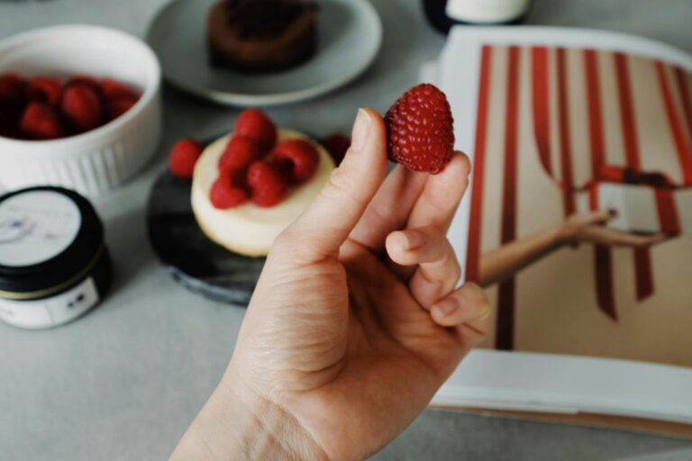 Close-up of a hand holding a raspberry with cheesecake and berries in the background. Perfect for food enthusiasts.