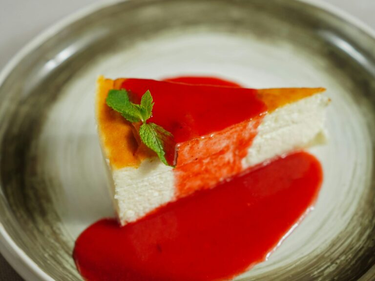 A slice of creamy cheesecake topped with strawberry sauce and fresh mint on a plate.