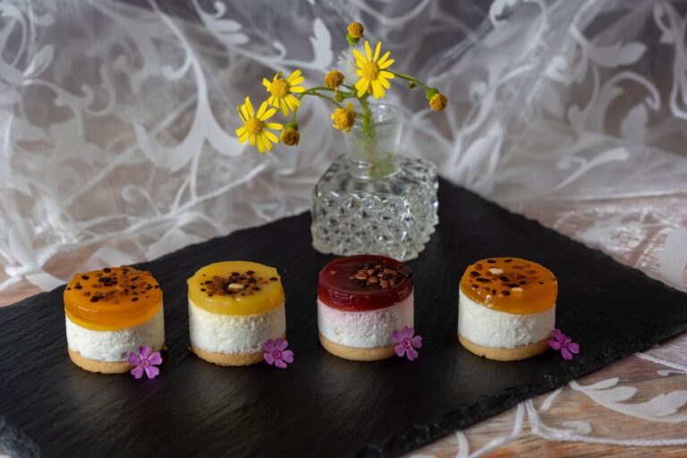 Assorted mini fruit cheesecakes with vibrant toppings presented on a slate board with flowers.
