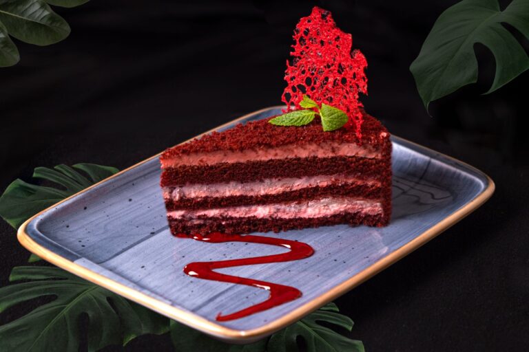 A close-up of a gourmet red velvet cheesecake slice on a stylish plate, featuring coral tuile and fresh mint garnish.