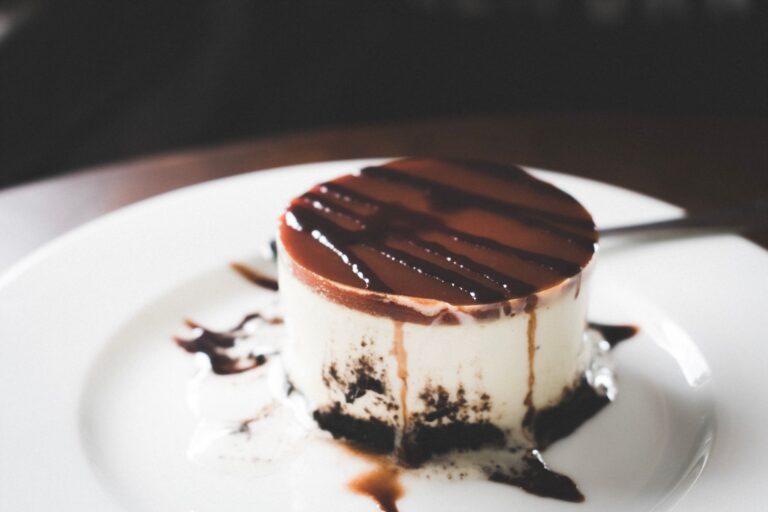 Close-up of a creamy cheesecake drizzled with rich chocolate syrup on a white plate.