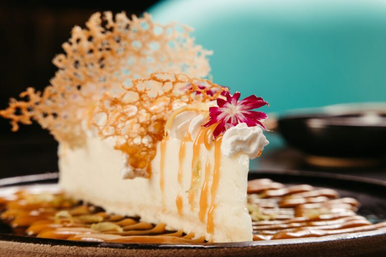 Close-up of gourmet cheesecake with caramel drizzle and edible flower garnishment on a decorative plate.