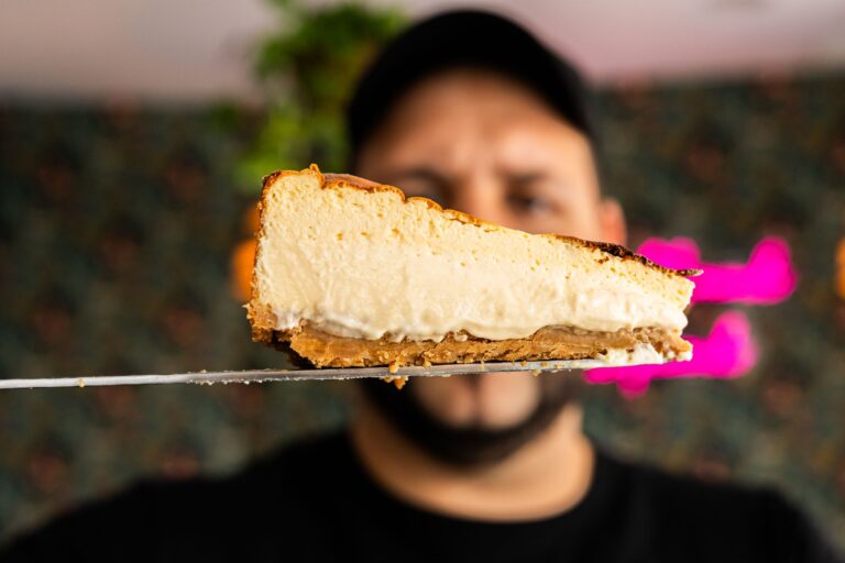 Close-up view of a creamy cheesecake slice held on a spatula, displayed against a blurred background.