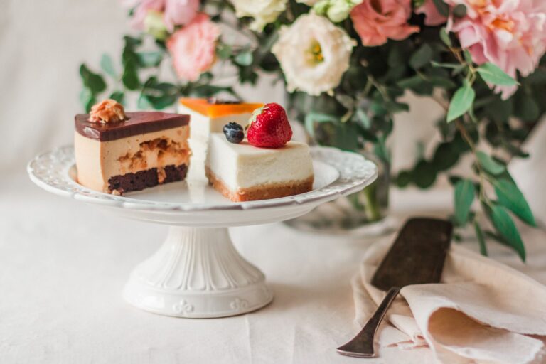 Appetizing pieces of cake covered with edible decorative elements and berries next to metallic kitchen spatula on dish towel and bouquet of flowers on blurred background