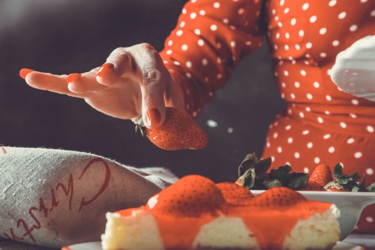 Woman in red polka dot dress placing fresh strawberry on cheesecake slice.