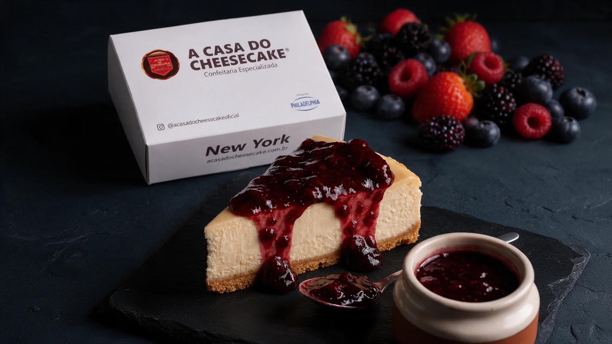Fatia de New York Cheesecake coberta com calda de frutas vermelhas sobre uma pedra escura, com um potinho de calda e a caixa da Casa do Cheesecake ao fundo